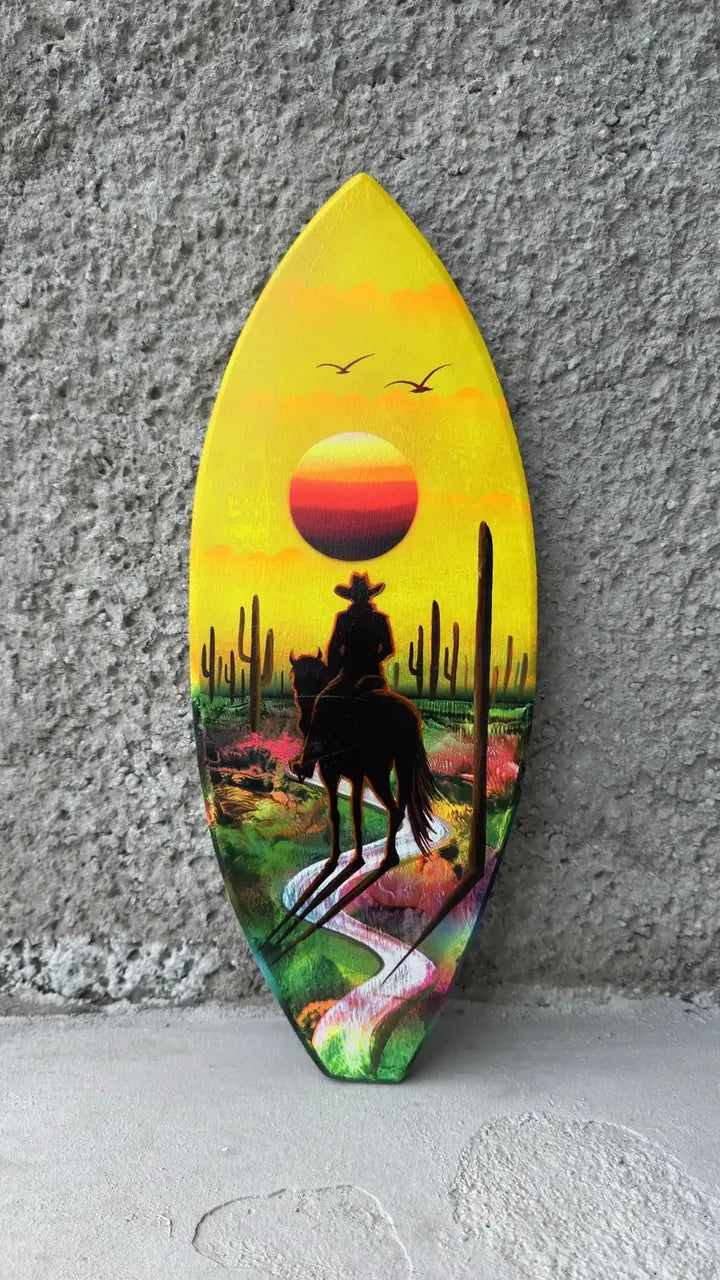 Surfboard-shaped painting of a rider on horseback in a desert at sunset