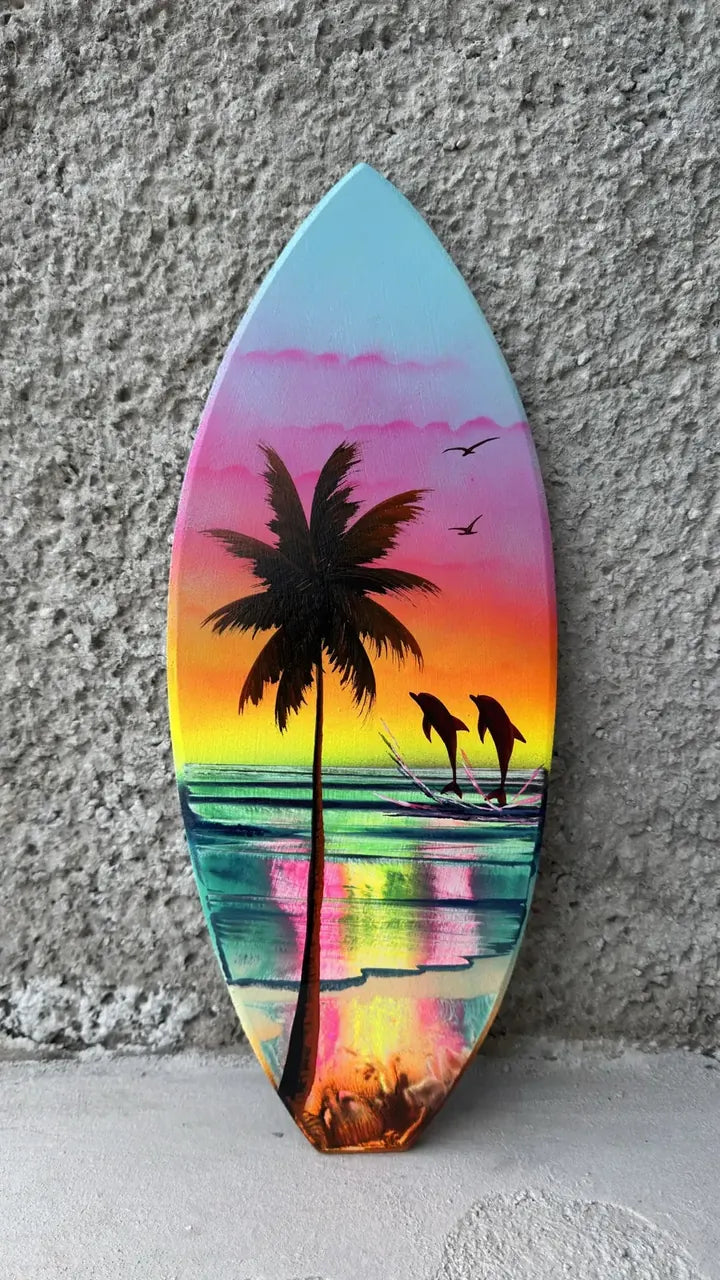 Surfboard-shaped painting of a palm tree and dolphins at a pink and yellow sunset